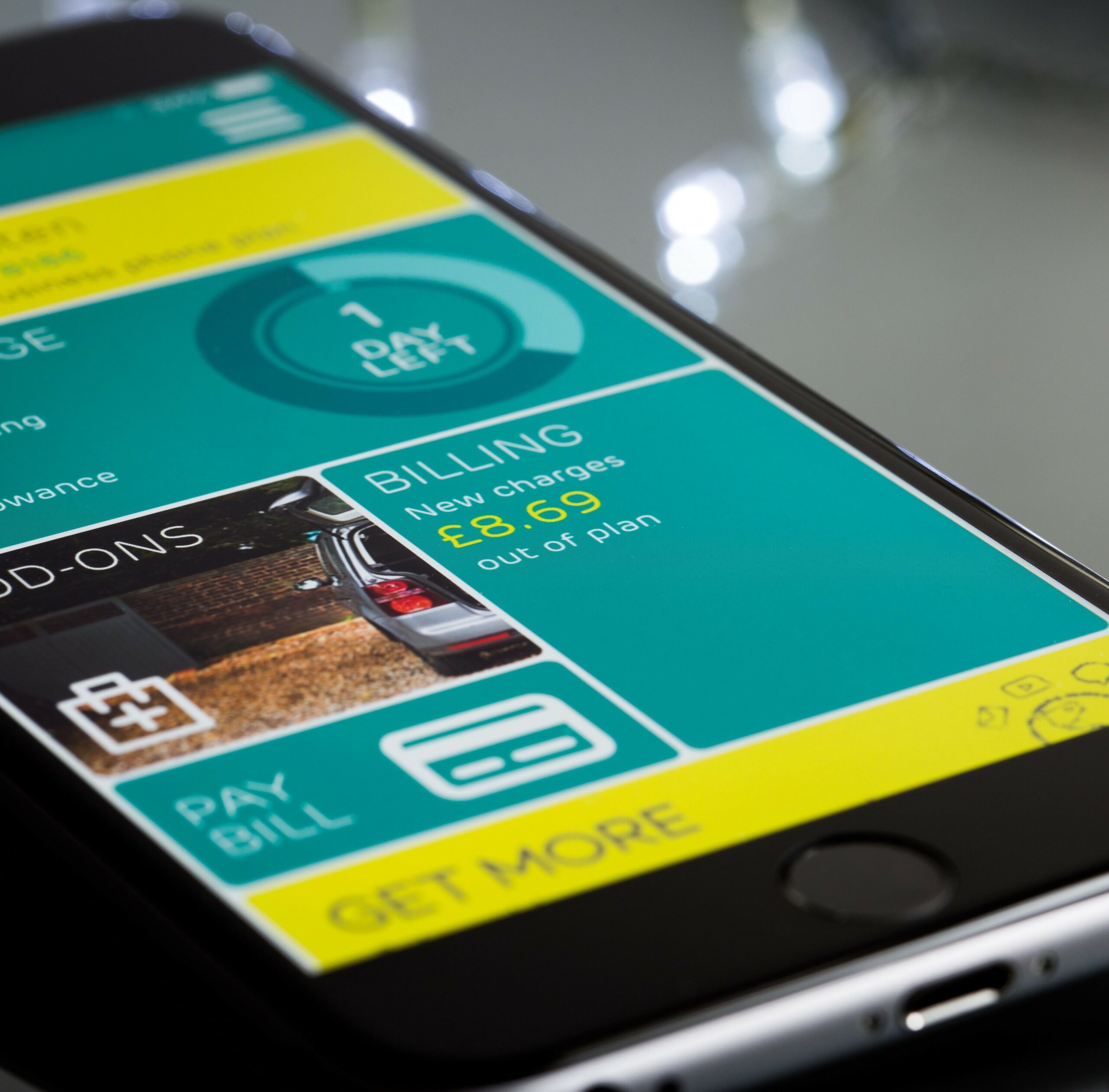 Vue en perspective d'un smartphone affichant une application mobile aux couleurs turquoise et jaune. L'écran présente un tableau de bord avec plusieurs sections, dont un encadré 'Billing' indiquant des frais hors forfait de £8.69, un compteur '1 day left', ainsi que des boutons pour payer sa facture et obtenir plus d'options.